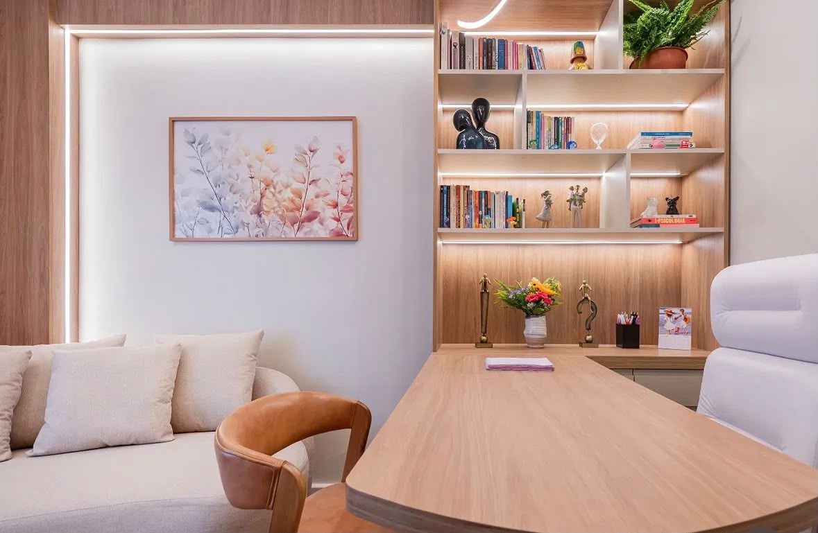 Sala de atendimento: sofá claro com almofadas, poltrona de couro, mesa de madeira, estante iluminada com livros e um quadro de flores.