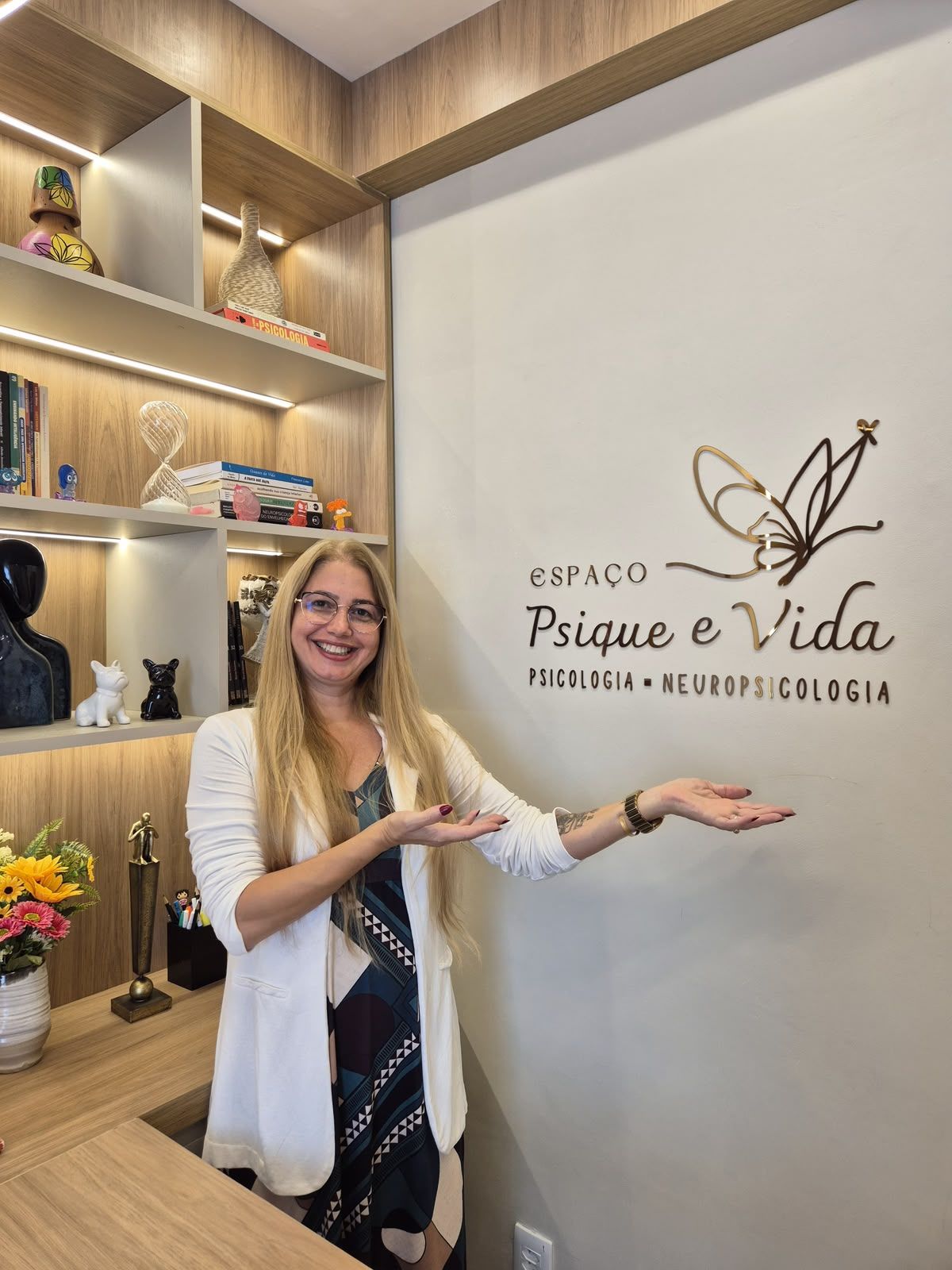 Viviane Gatis em pé no consultório, apontando para o letreiro do Espaço Psique e Vida na parede.