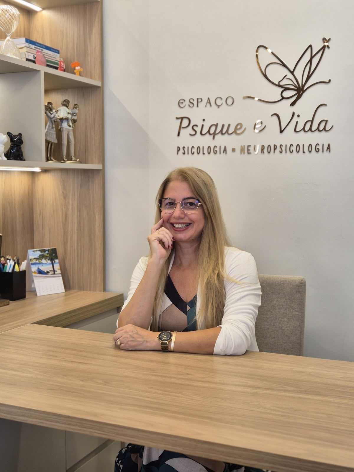Viviane Gatis à mesa do consultório, sorrindo, com o letreiro do espaço na parede ao fundo.