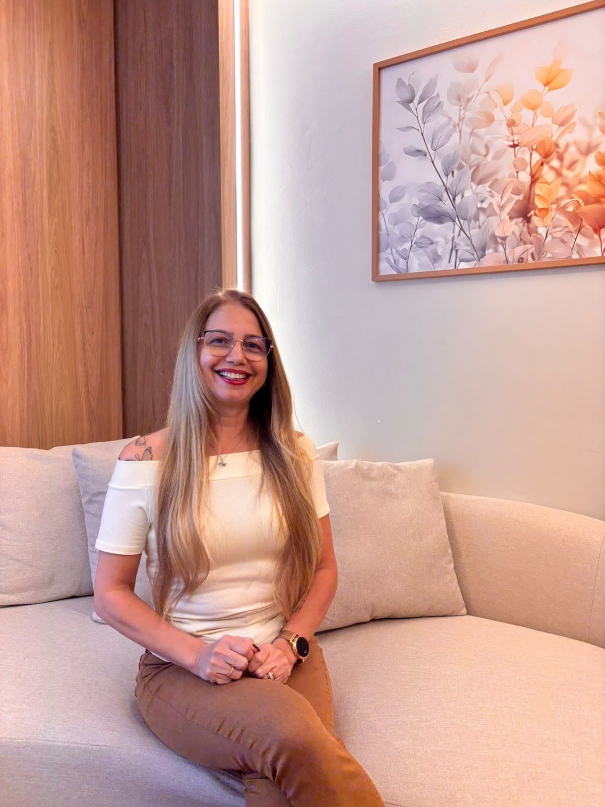 Viviane Gatis sorrindo, sentada no sofá do consultório, com um quadro de folhas ao fundo.
